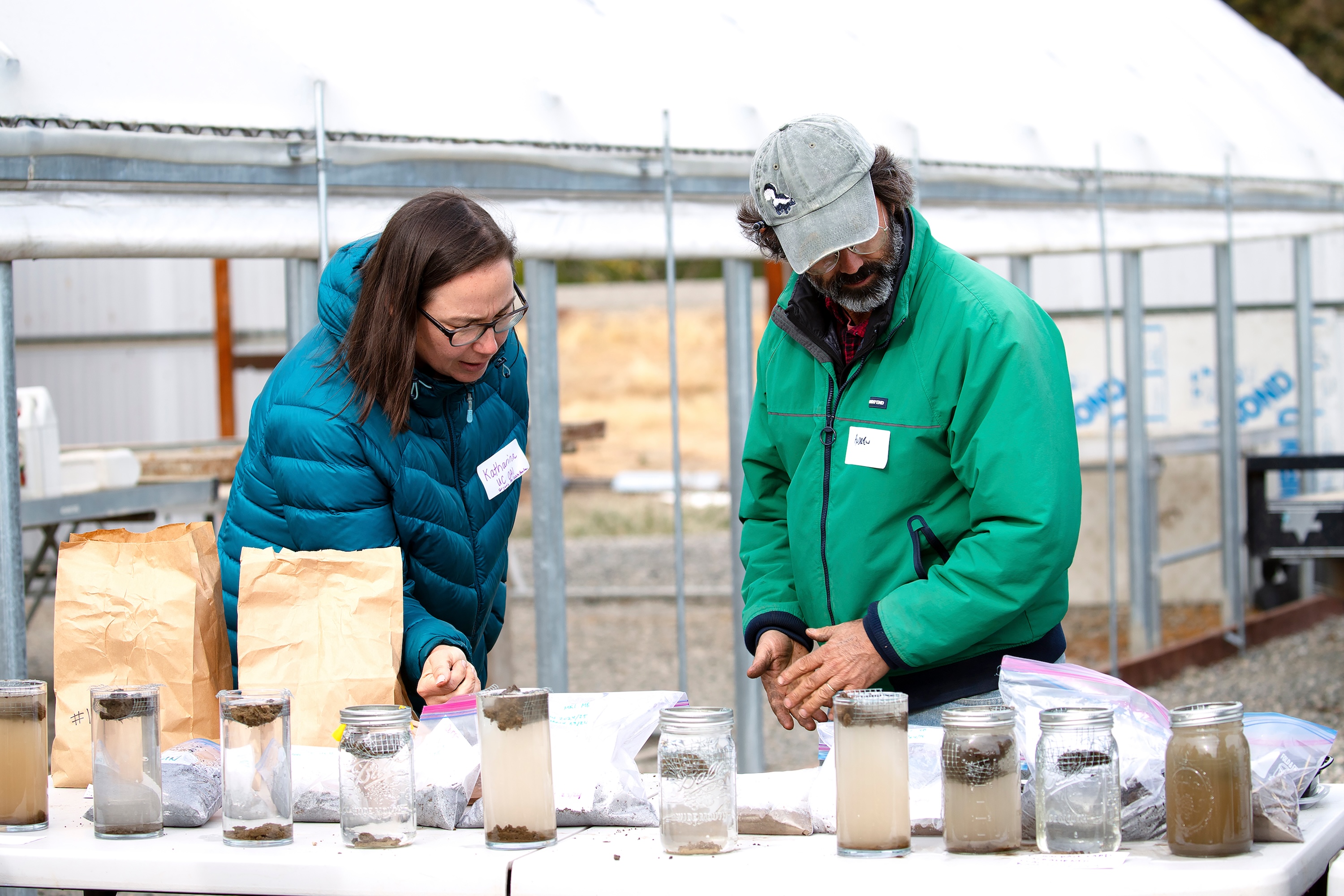 A woman and a blue puffy jacket and a bearded man in a green jacket look at slake test results in glass jars.