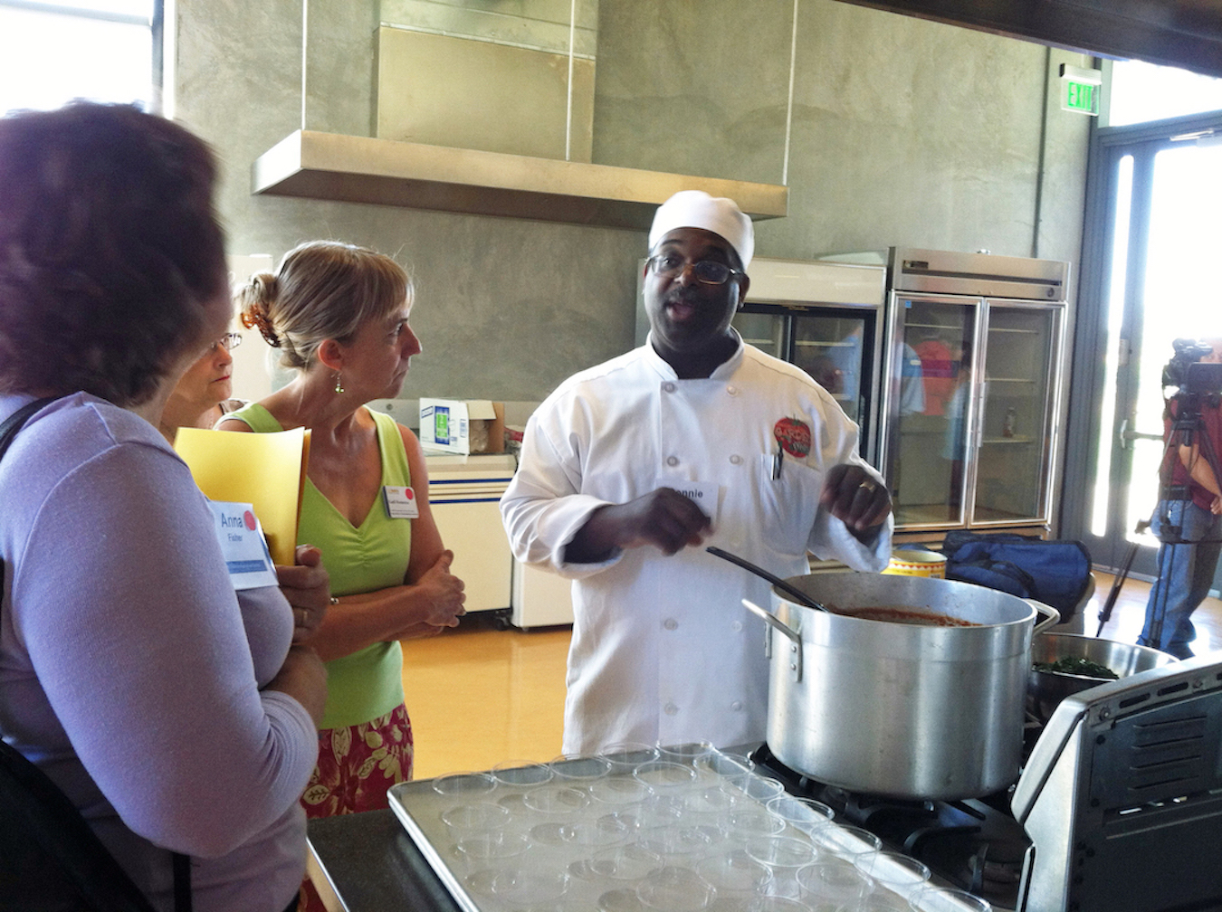 Two people listen as a chef in white hat and uniform speaks while stirring large pot of soup.