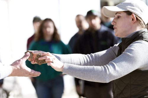 A woman with a baseball cap on hand soil pieces to an person with their hand held out while several others look on. 